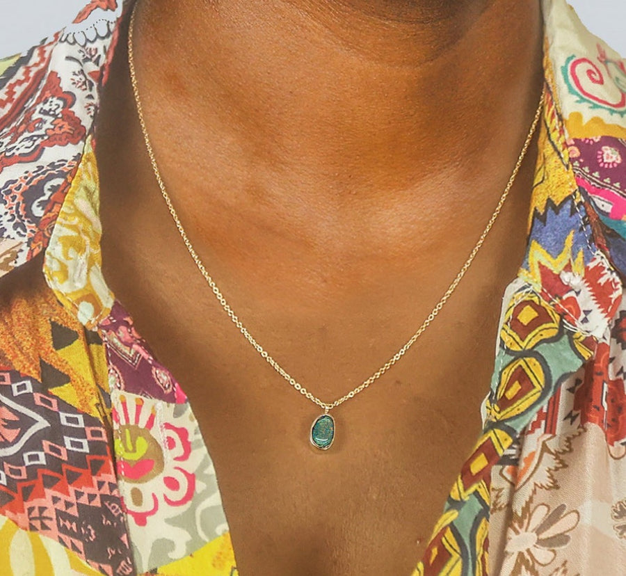 A blue and gold opal in a round setting, hanging from a thin gold chain worn around a woman's neck. 